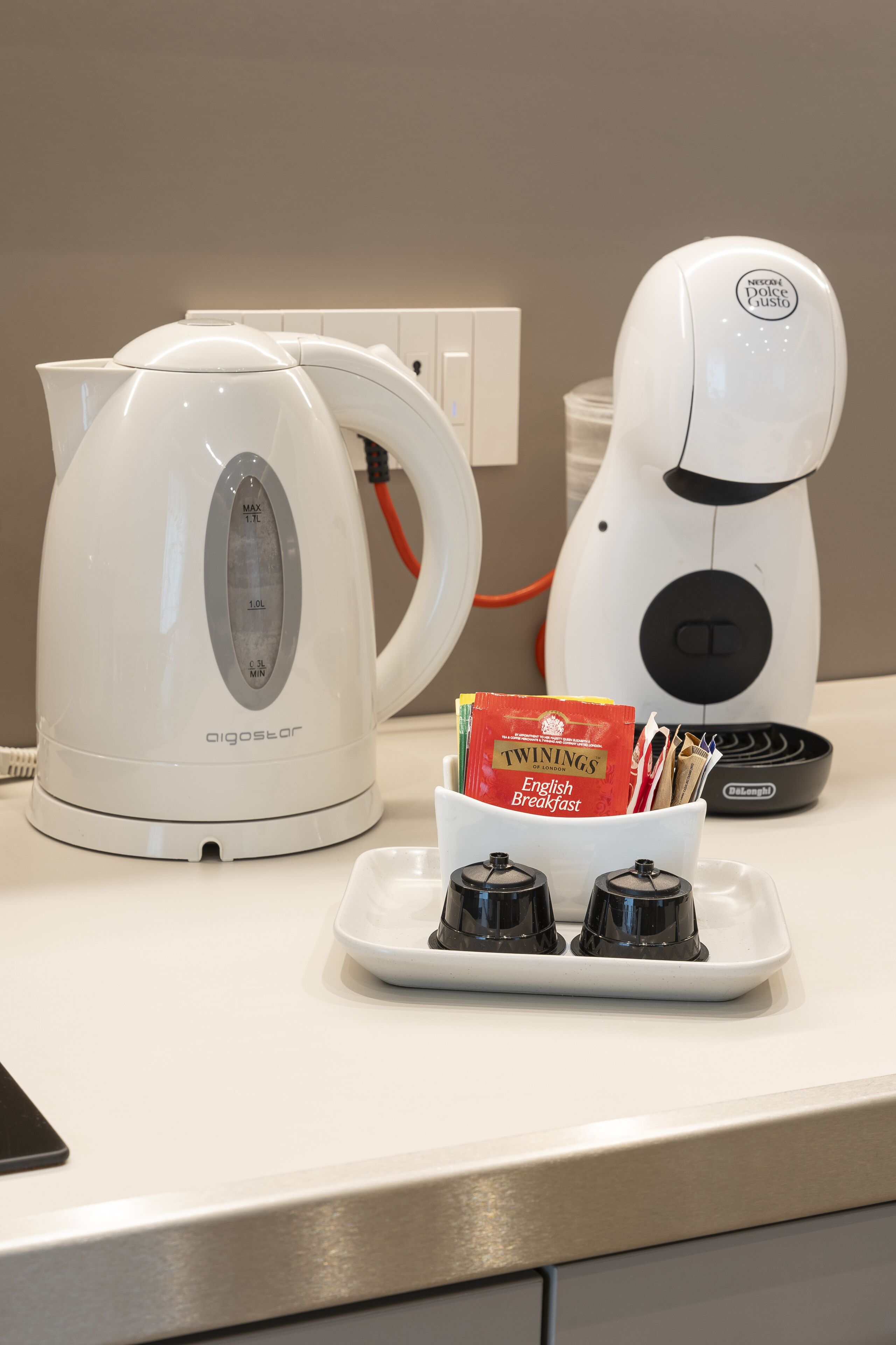 A kitchen counter with a white electric kettle, a coffee maker, and a tray with a packet of Twinings English Breakfast tea and condiments.