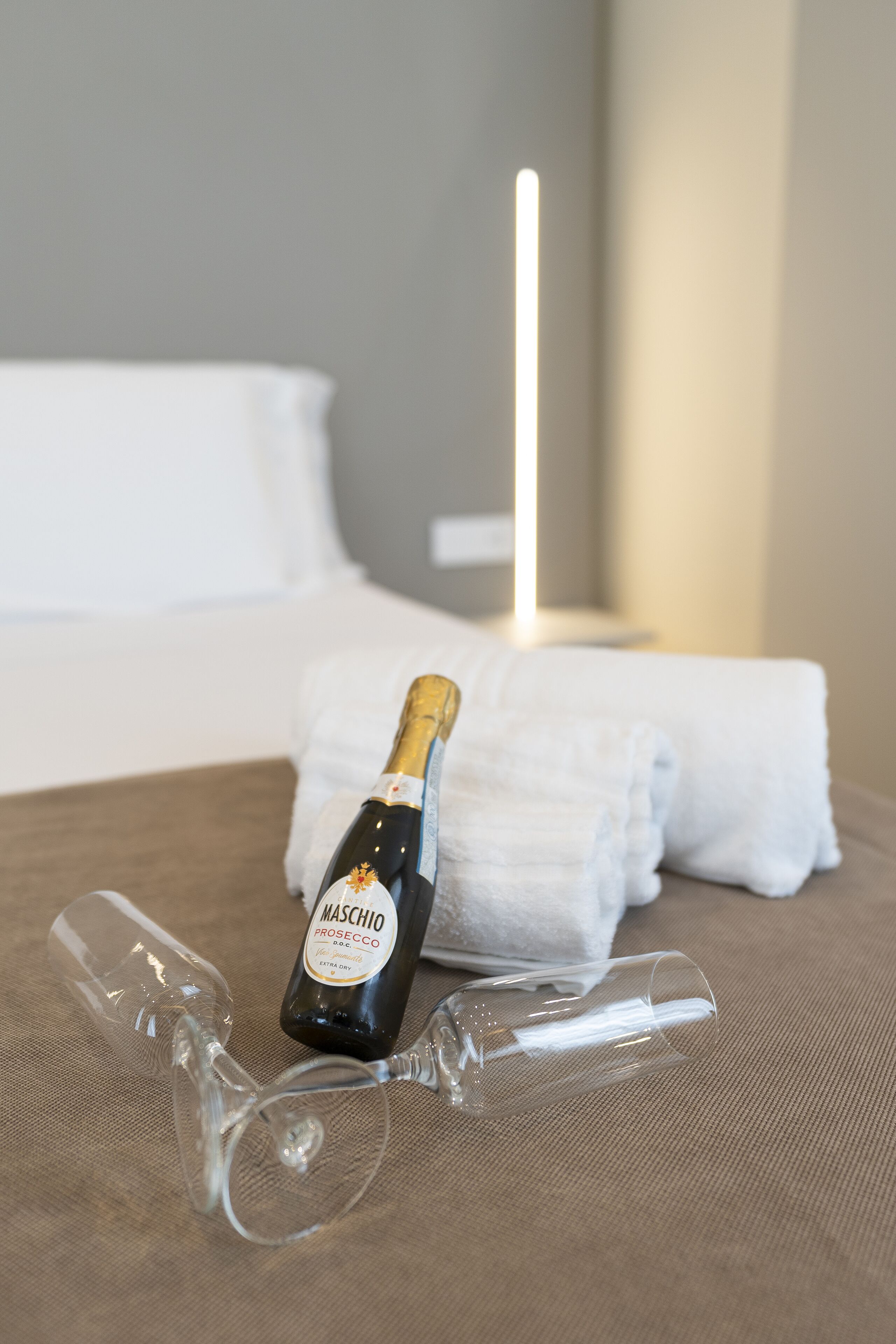 A bottle of Maschio Prosecco and two wine glasses on a bed with white linens and a visible headboard.