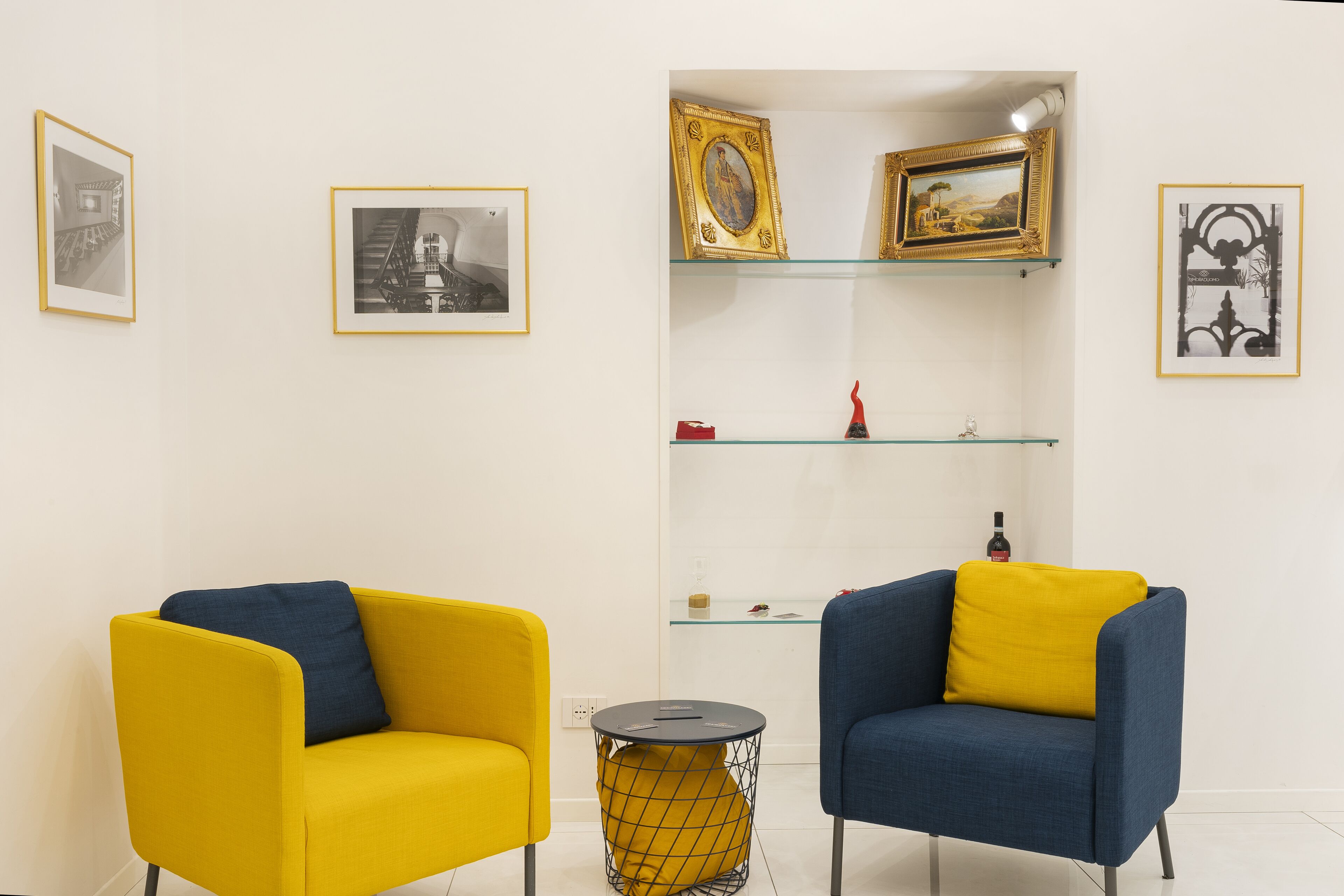 A room with two armchairs, one yellow and one blue, with cushions. There are framed pictures on the walls and a glass shelf with decorative items.