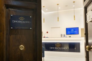 Un'area reception con una porta in legno con su scritto “DIMORADUOMO NAPOLI” e una scrivania reception con un monitor che mostra lo stesso logo.
