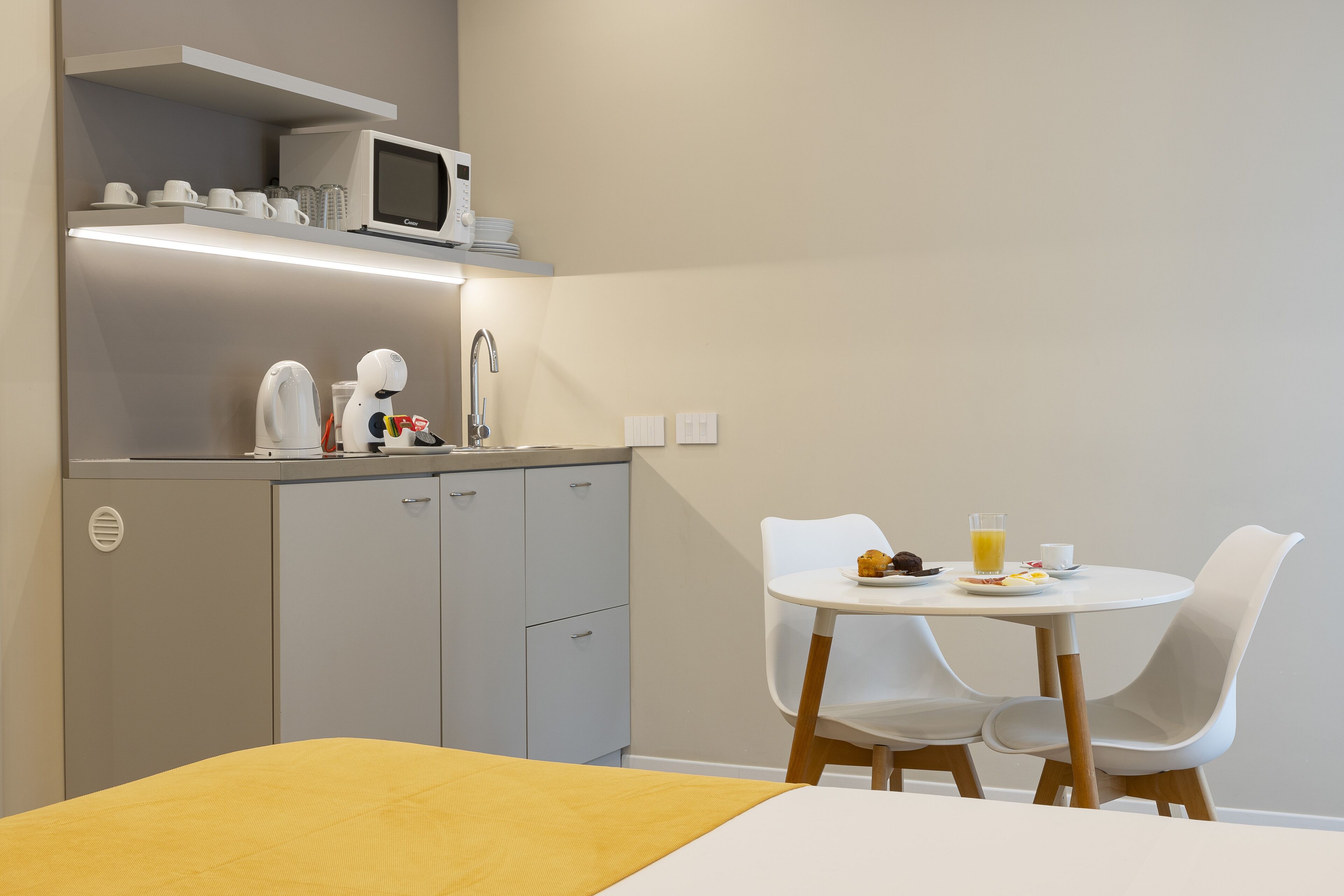 A compact hotel room with a bed, a small kitchenette, a dining table with chairs, and a microwave on the shelf.