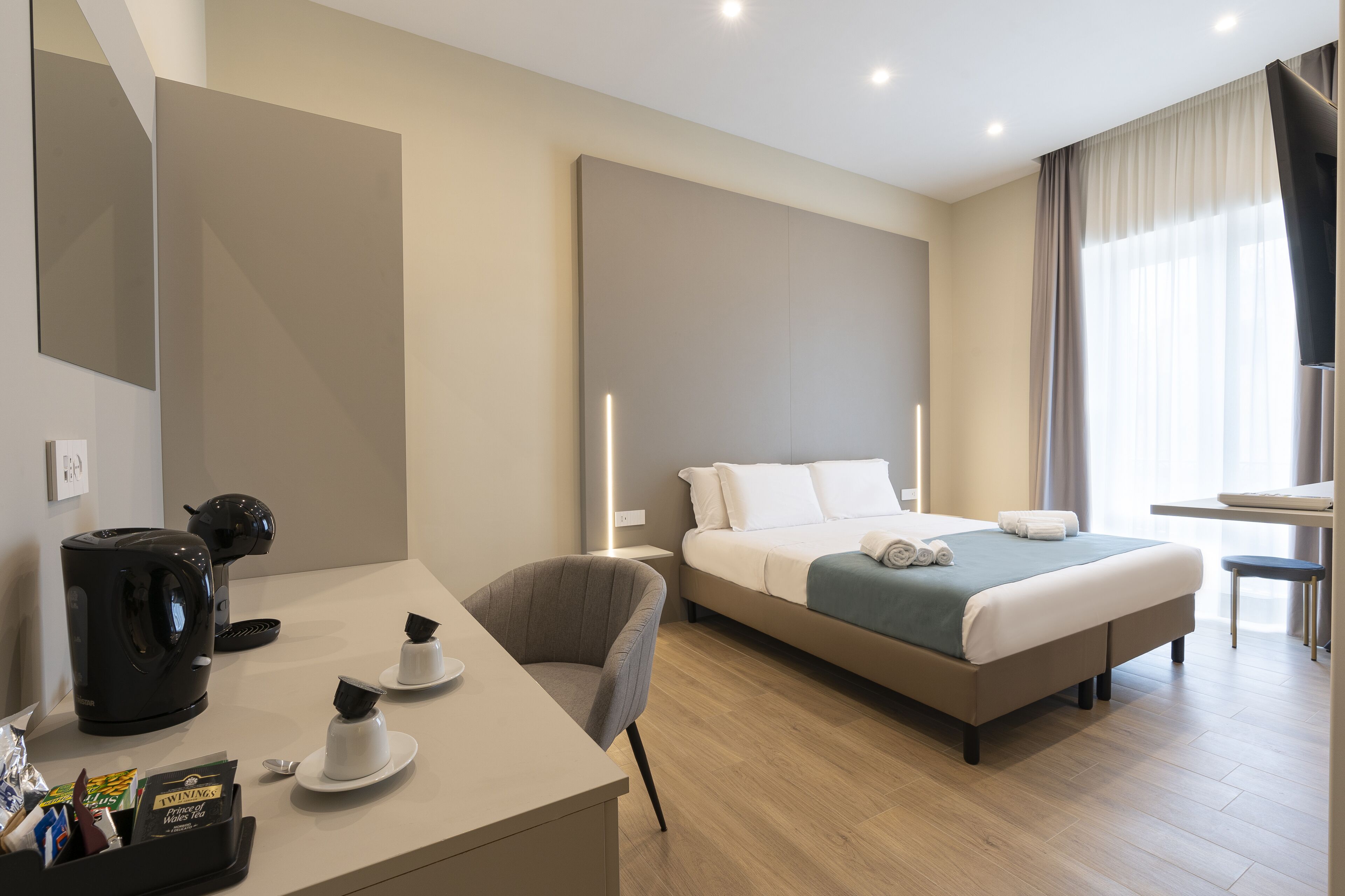 Ein modernes Hotelzimmer mit einem Bett, einem Schreibtisch mit Kaffeemaschine, einem Sessel und einem kleinen Tisch mit Tassen.