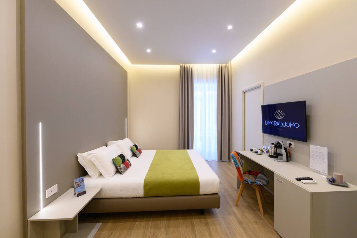 A modern hotel room with a bed, a desk, a chair, a television, and a window with curtains.
