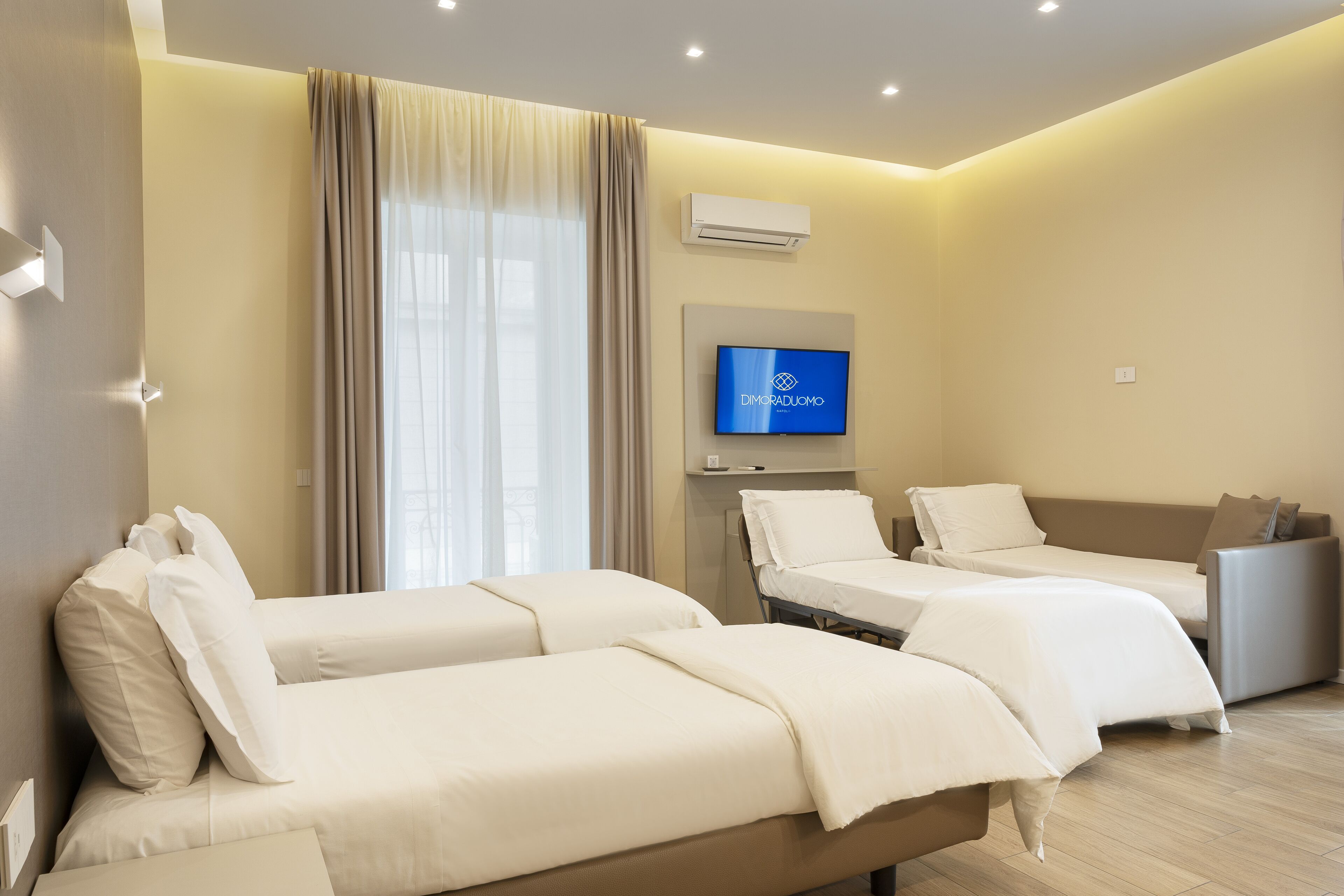 Ein Hotelzimmer mit zwei Einzelbetten, einem Schlafsofa, einem Flachbildfernseher und einer Klimaanlage.