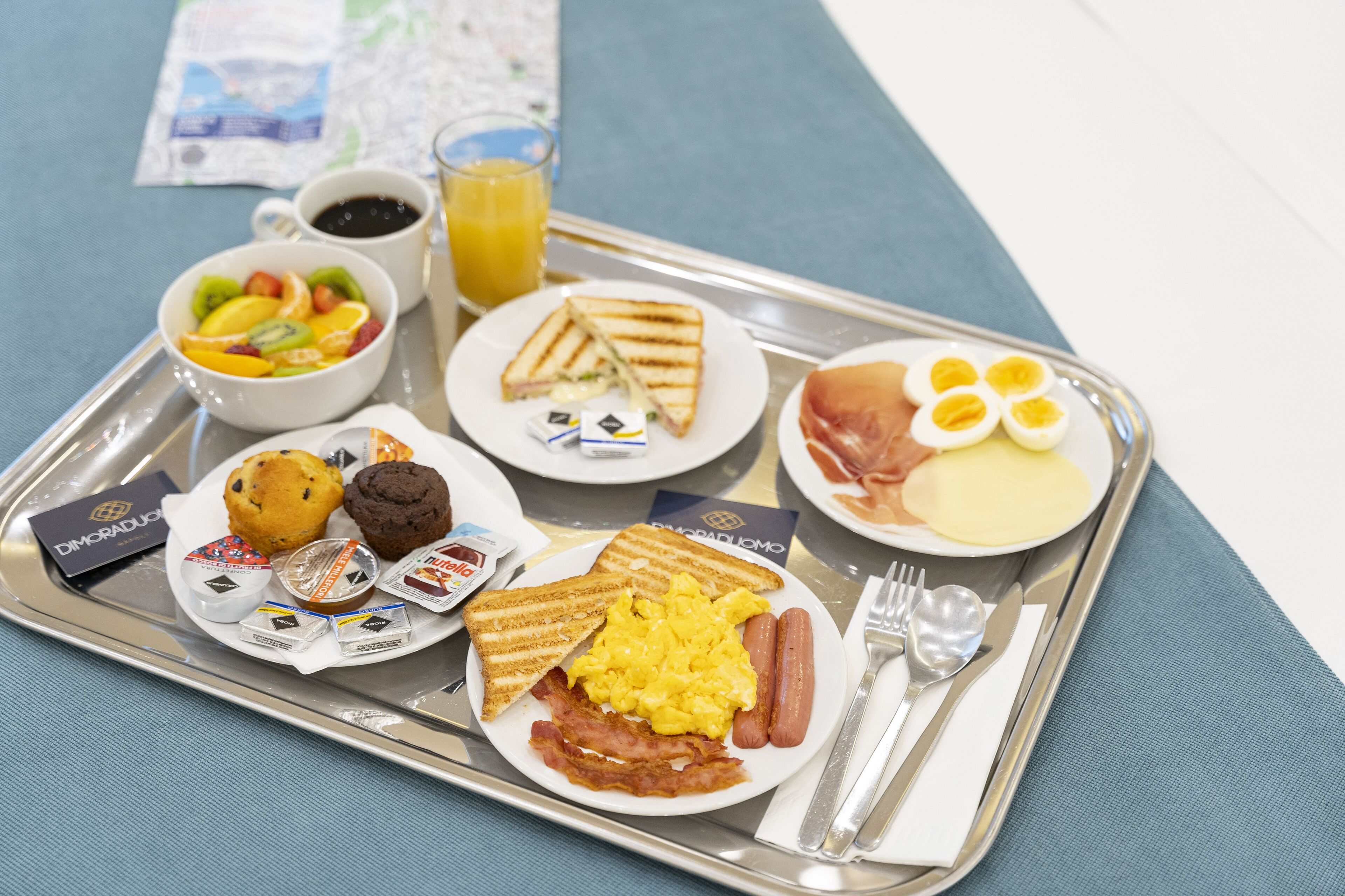 Bandeja de café da manhã com torradas, ovos, bacon, linguiça, muffins e uma saladeira de frutas.