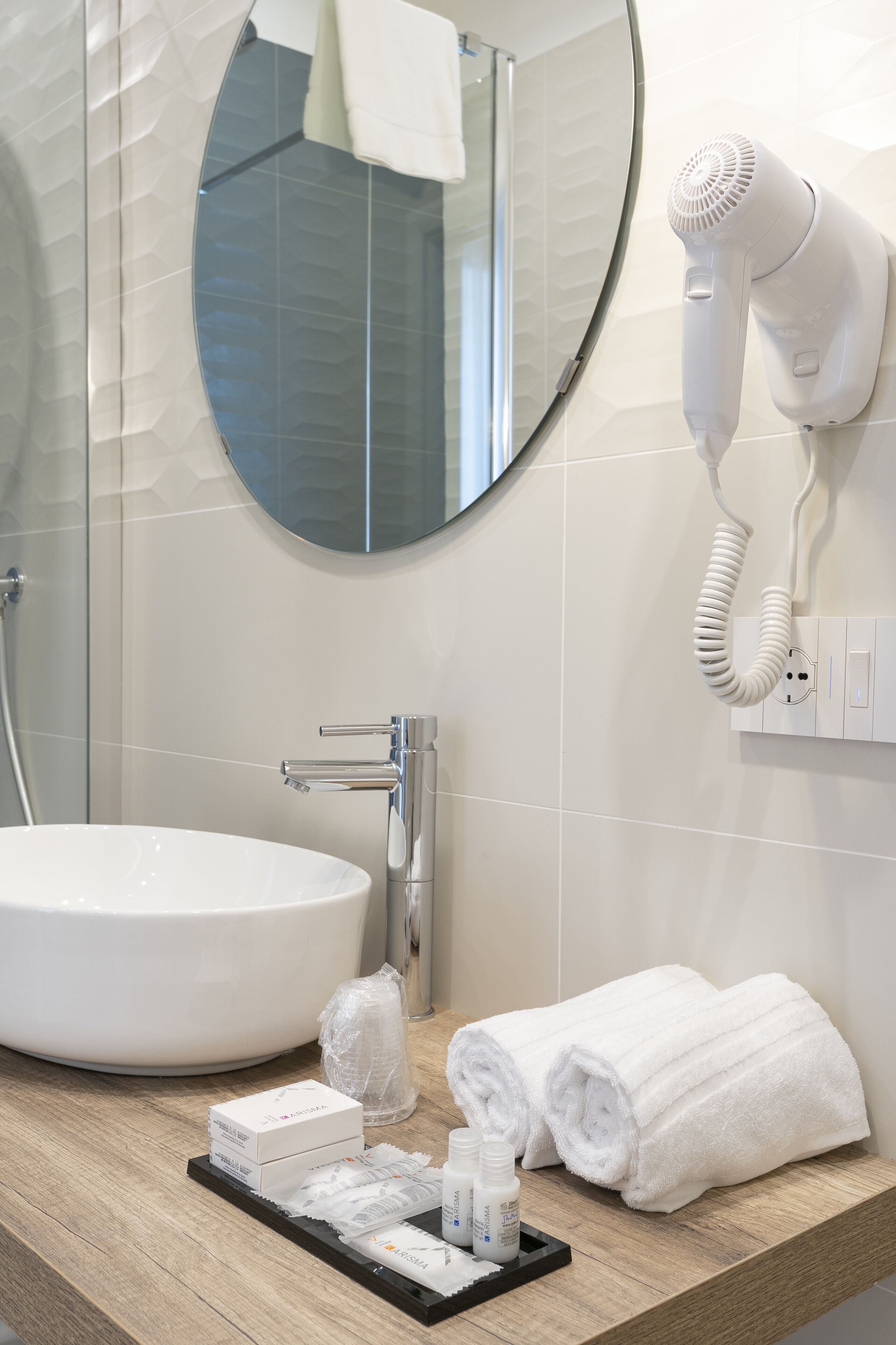 Un baño con un lavabo blanco, un espejo redondo, un secador de pelo montado en la pared y toallas ordenadas sobre el mostrador.