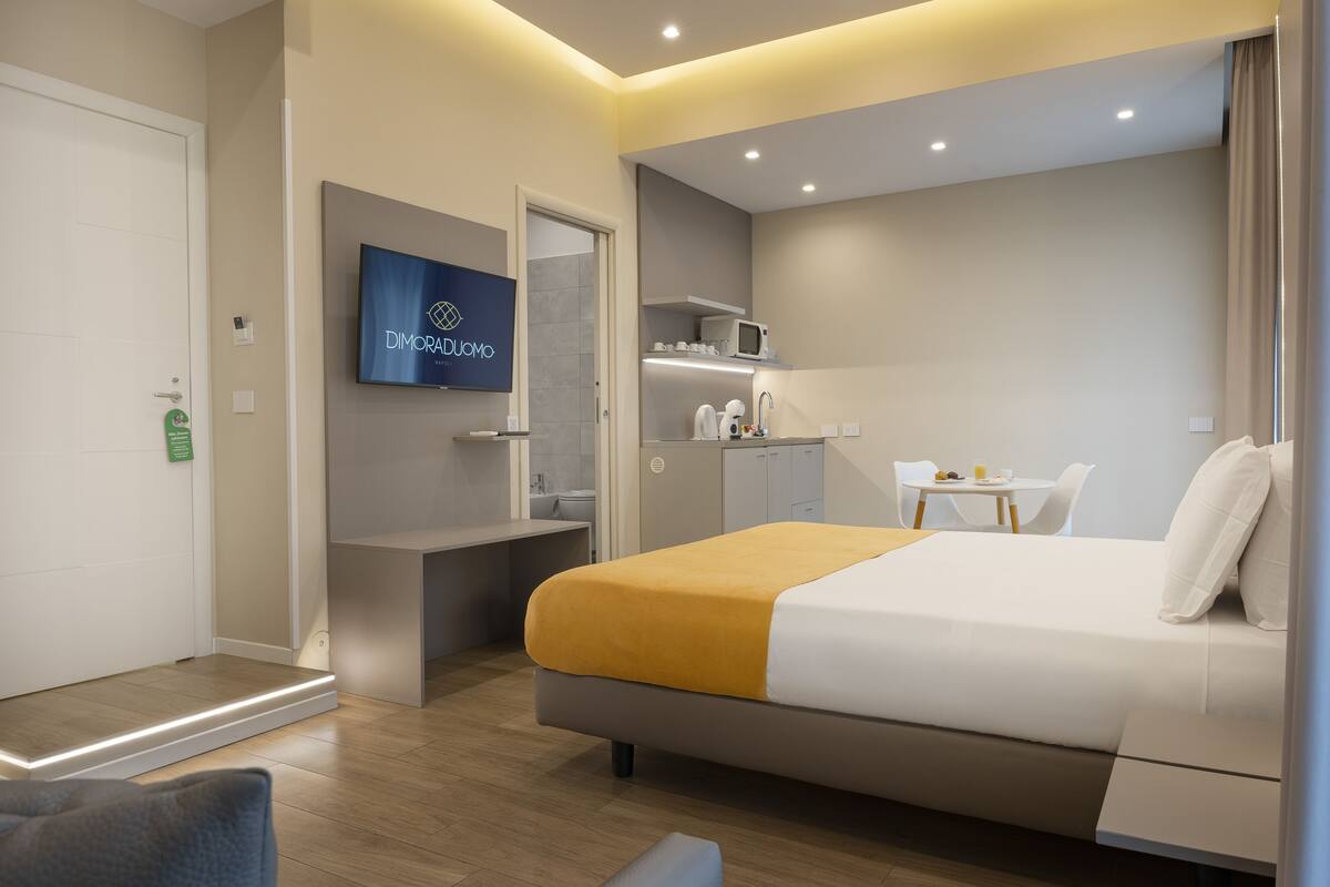 A modern hotel room with a bed, a desk, a TV, and a small kitchenette.