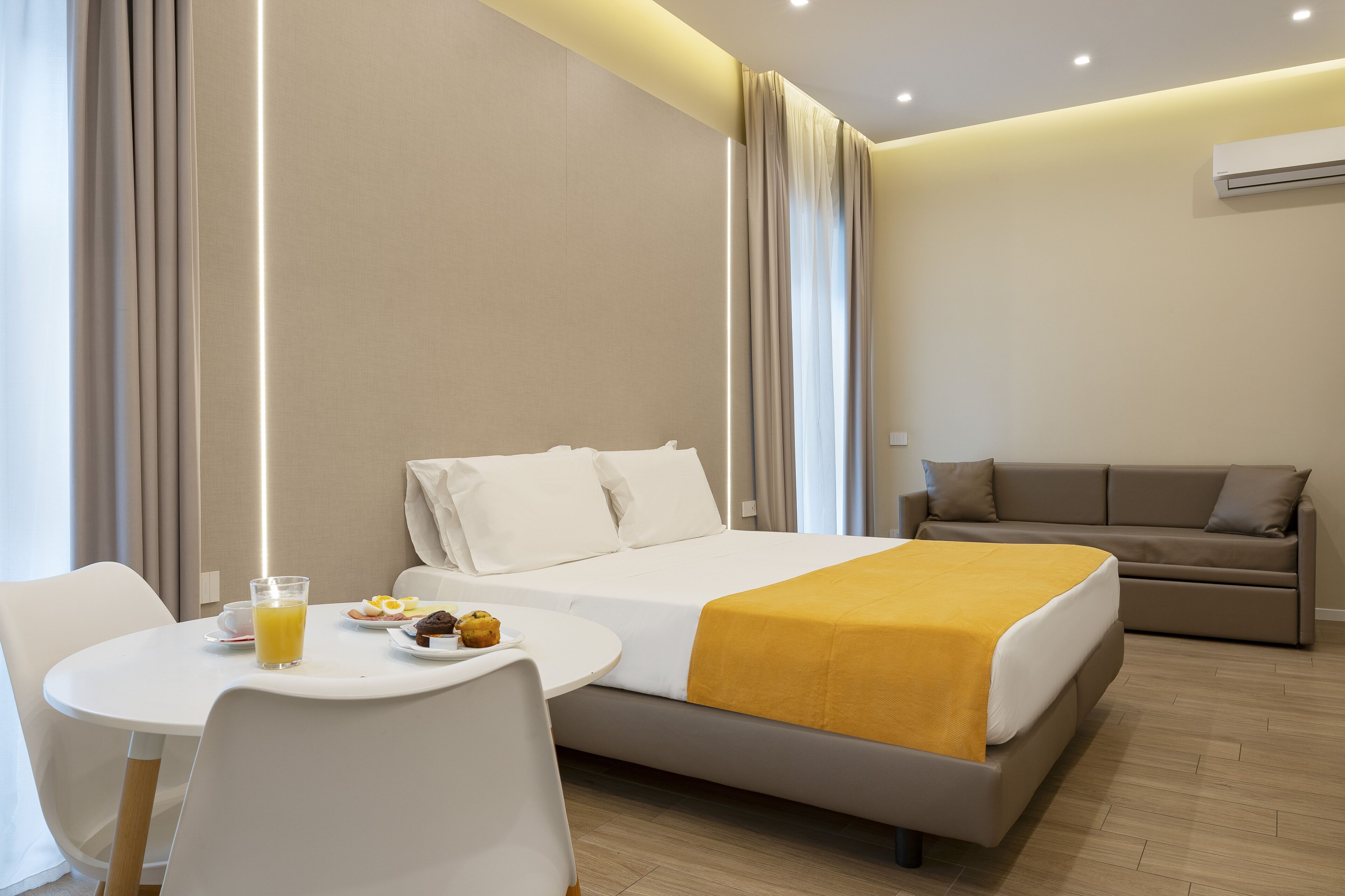Une chambre d’hôtel avec un lit, un canapé, une table à manger où le petit-déjeuner est servi, et une fenêtre avec des rideaux.