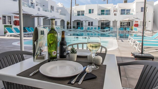 A table set for two with a bottle of wine, a glass of white wine, and a plate, overlooking a pool area with lounge chairs and palm trees. - Guinate Club Apartamentos (Tías)