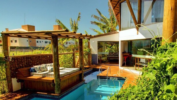 Un área de piscina moderna al aire libre con terraza de madera, asientos y una sala con paredes de vidrio.