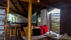 Una habitación acogedora con techo de madera, una cama con almohadas de colores, un escritorio pequeño con una computadora y una ventana con vistas al verdor.