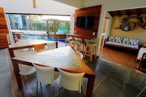 Comedor con una mesa grande y sillas, un televisor montado en la pared y una piscina visible a través de puertas de vidrio.