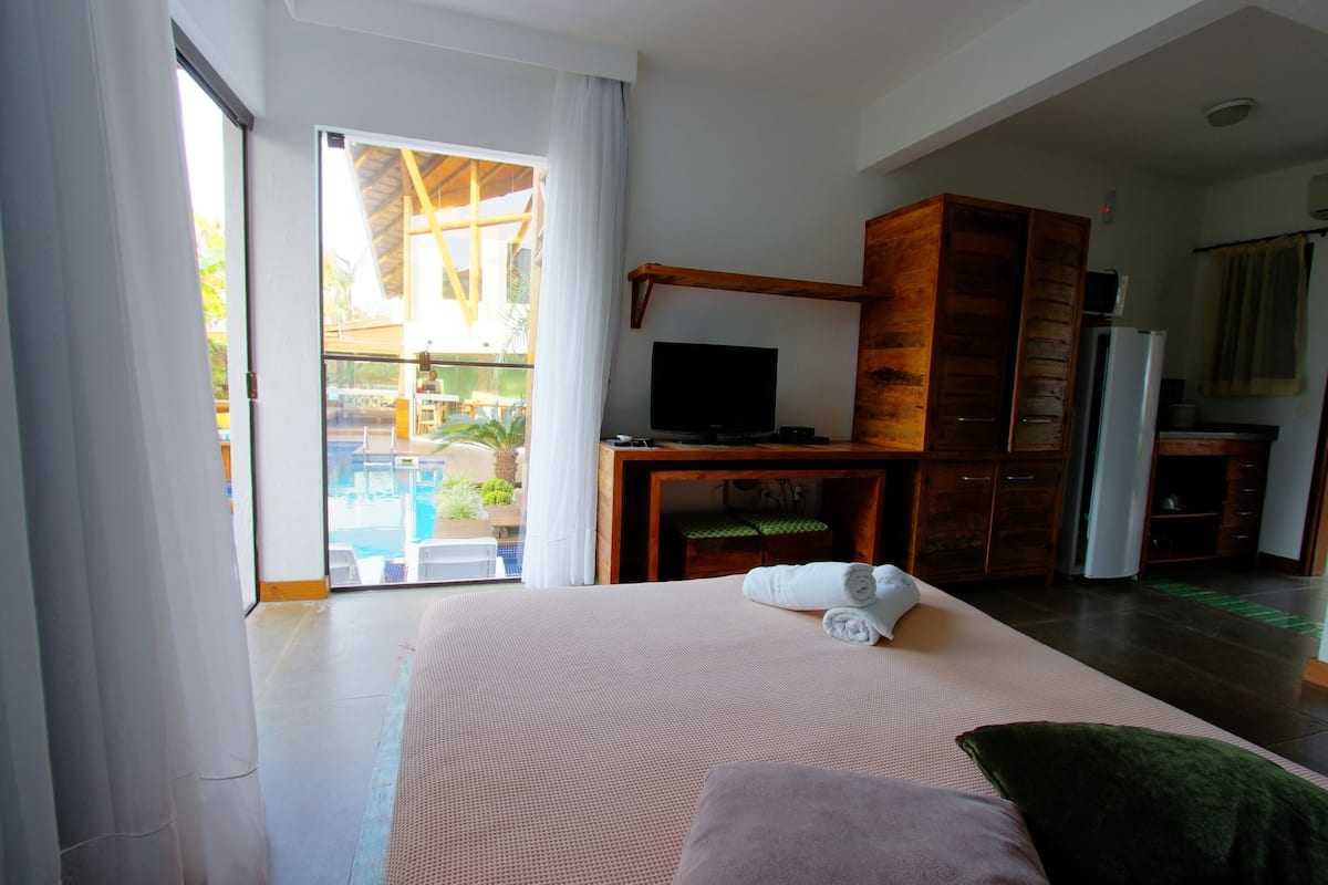 Una habitación de hotel con una cama grande, televisión, armario de madera y puerta corrediza de vidrio que da a una zona de piscina.