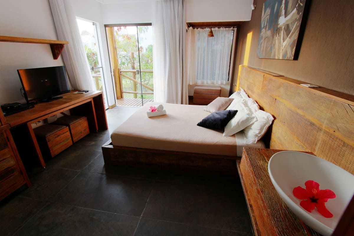 Un dormitorio con cama, escritorio, televisor y baño con una flor, además de vistas al exterior.