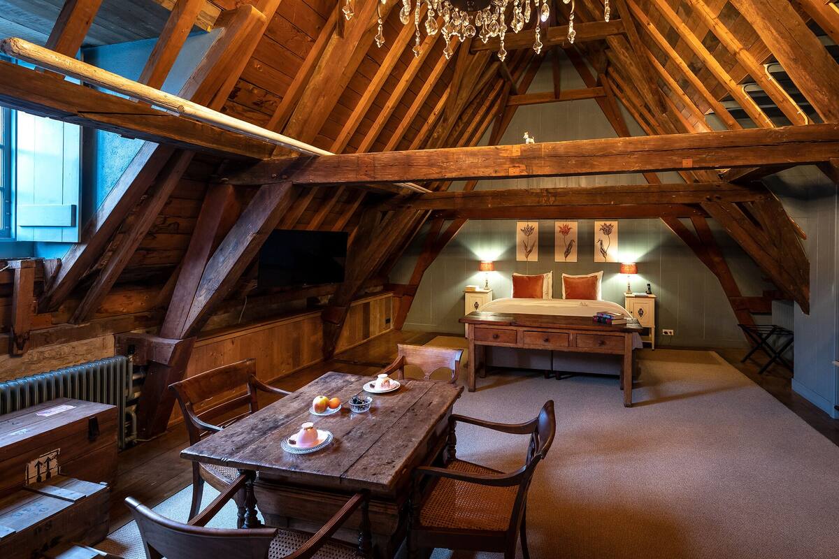 Une chambre mansardée au style rustique, avec des poutres apparentes, une table à manger en bois et des chaises, un lit avec une table de chevet et des lampes fixées au mur.