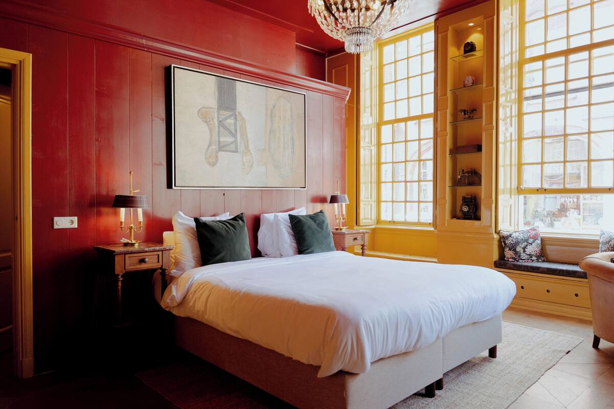 Une chambre spacieuse avec un grand lit, deux tables de chevet avec des lampes, une fenêtre donnant sur un beau paysage et un lustre.
