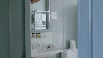 Een badkamer met een witte wasbak, spiegel en handdoekhouder. Er is een toilet met een witte tank en een prullenbak. De muren zijn lichtblauw en er is een houten balk aan het plafond.