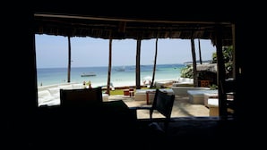 Vista de una playa desde un área de estar al aire libre cubierta, con techo de paja, asientos blancos acolchados y vista al océano con barcos.