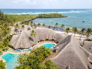A resort with a pool surrounded by thatched-roof structures, palm trees, and a view of the ocean. - Blue Bay Village (Watamu)