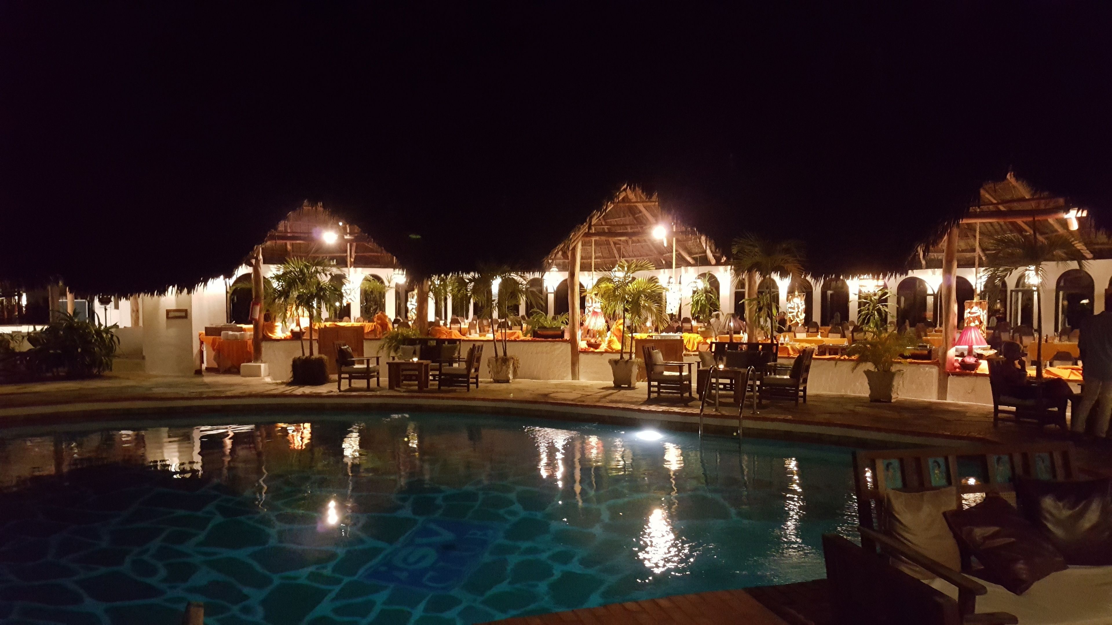A nighttime pool area with multiple dining tables and chairs, surrounded by palm trees and illuminated by ambient lighting.