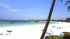 Una playa con arena blanca, agua turquesa cristalina y varios barcos. Se ven palmeras y sillas de playa.