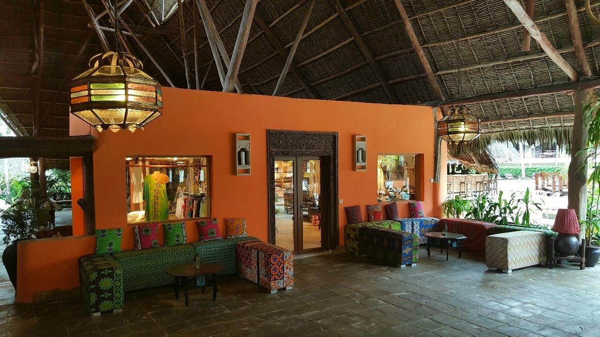 Un espacio interior con sofás y mesas coloridos, techo de paja y acceso visible a una tienda.