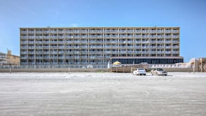 A large, multi-story building with numerous balconies, a parking area with several cars, and a clear sky.