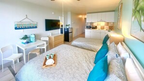 A compact hotel room with a bed, a desk with a chair, a kitchenette, and a ceiling fan.