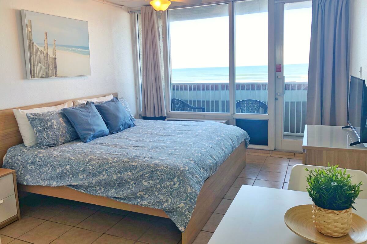 A hotel room with a bed, a TV, a balcony with a view of the sea, and a ceiling fan.