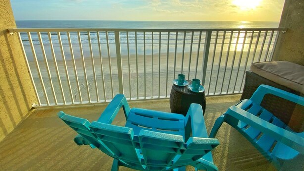 A balcony with a view of the ocean and a sunset. There are two blue chairs and a small table with two cups.