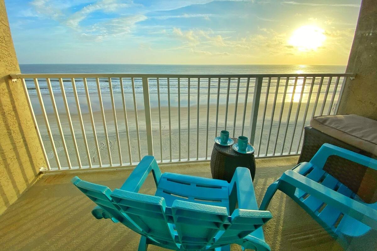 A balcony with a view of the ocean and a sunset. There are two blue chairs and a small table with two cups.