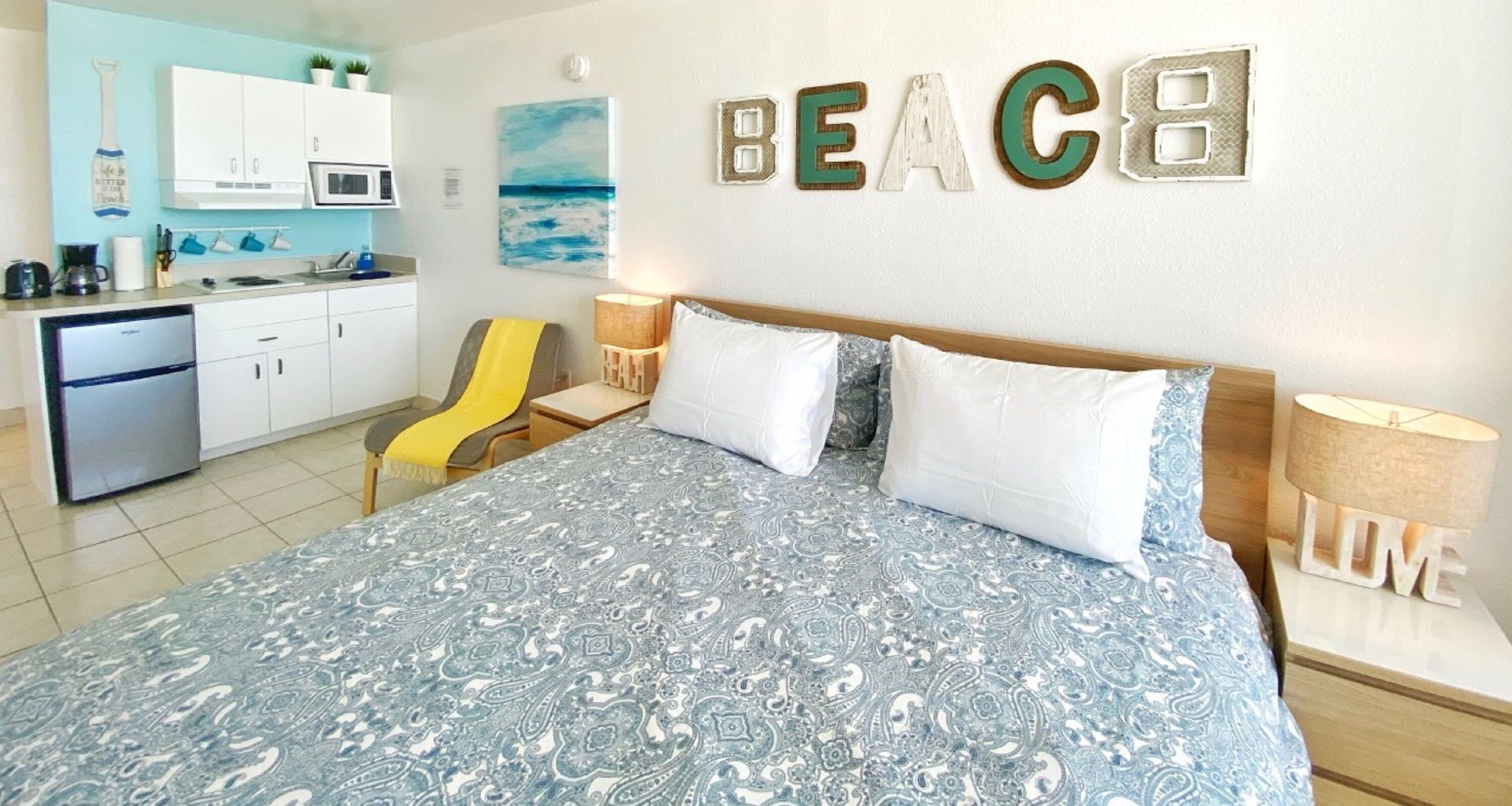 Un dormitorio con una cama, dos almohadas, una mesita de noche y una silla. Hay una zona de cocina con armarios y un microondas. En la pared se muestra la palabra 'BEACH'.