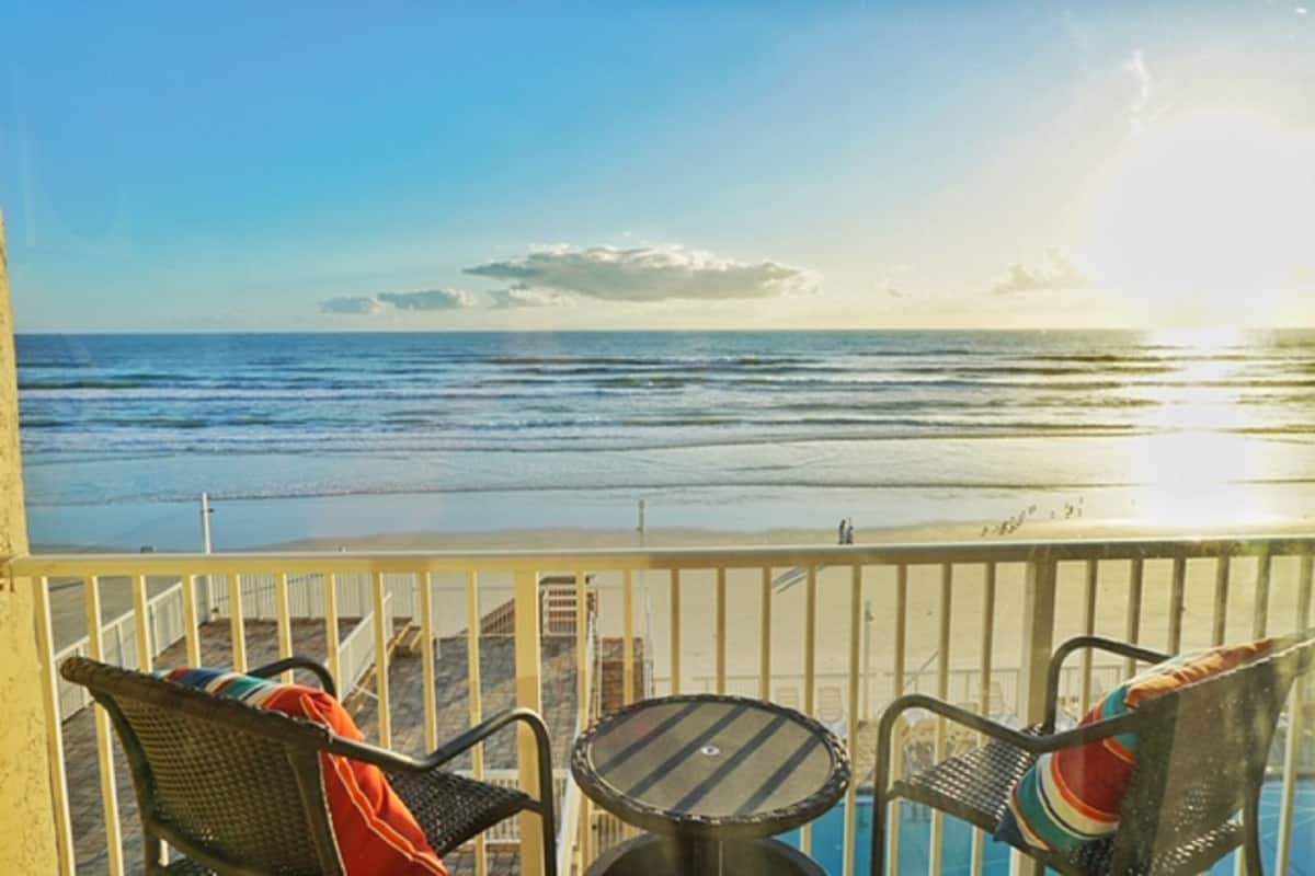 A balcony with two wicker chairs and a small table, overlooking a beach with the ocean and a clear sky.