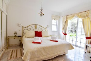 A spacious bedroom with a large bed, a chandelier, and a view of greenery through the window.