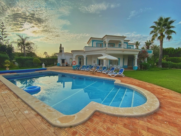 Una piscina con sillas de descanso, un porche cubierto y una casa grande con jardín.