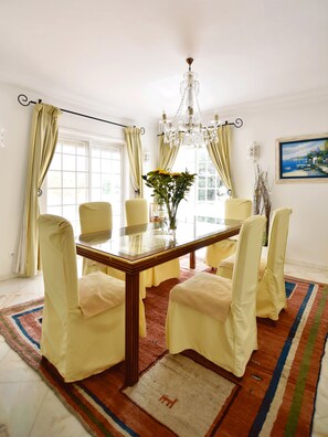 A dining room with a wooden table, six chairs, a chandelier, and a large window with curtains.