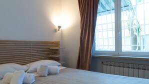 Uma cama bem arrumada com lençóis e toalhas brancas, uma cabeceira de madeira, uma luminária fixada na parede e uma janela com cortinas.