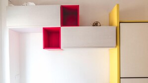 Een compacte kamer met een witte tafel en twee stoelen, een klein rode plankkast, en een gele muur.