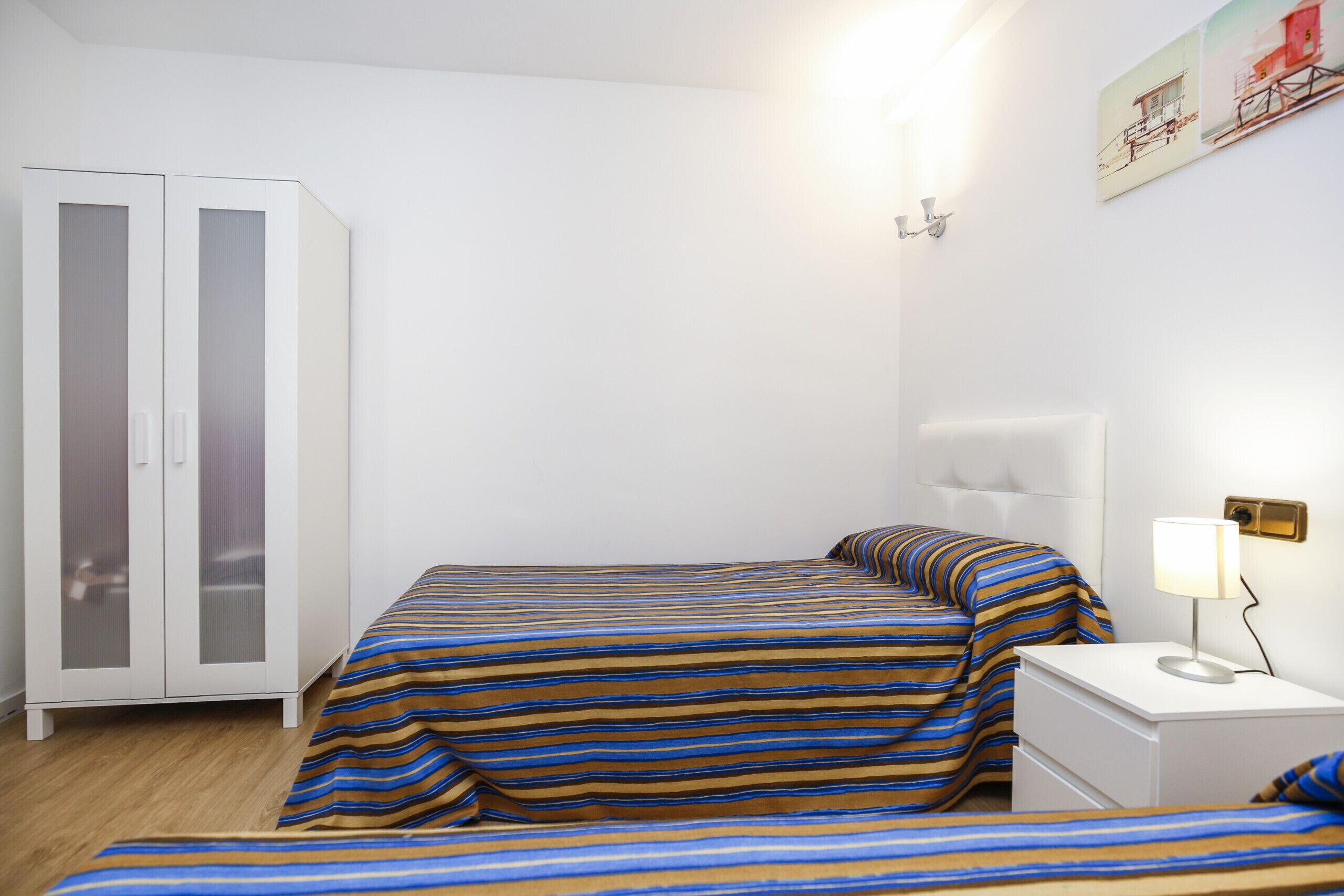 A bedroom with a bed, a nightstand, a lamp, a wardrobe, and a wall-mounted light fixture.