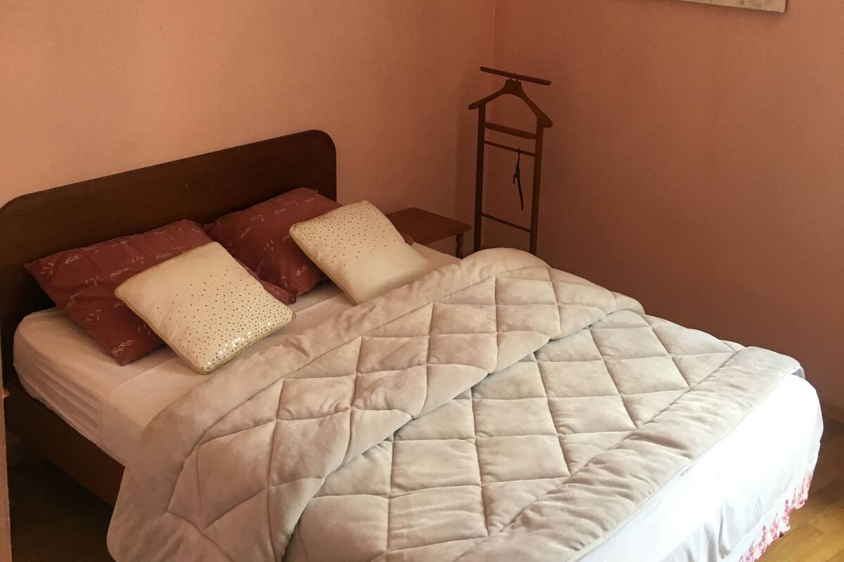 Un dormitorio con cama, una lámpara colgante, una mesita de noche pequeña y un adorno en la pared.