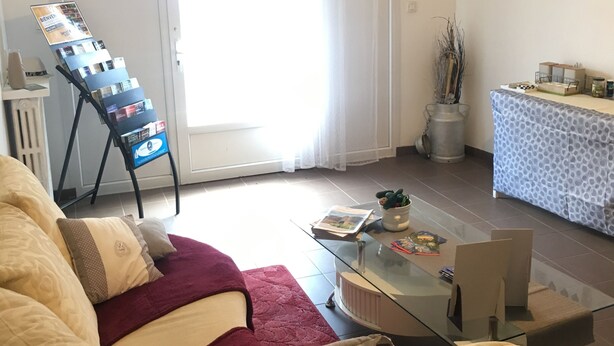 A living room with a sofa, a glass coffee table, a small table with a plant, a magazine rack, and a sliding glass door leading to a balcony. - Le Palmyre Soleil de l'Amazonie (La Douze)