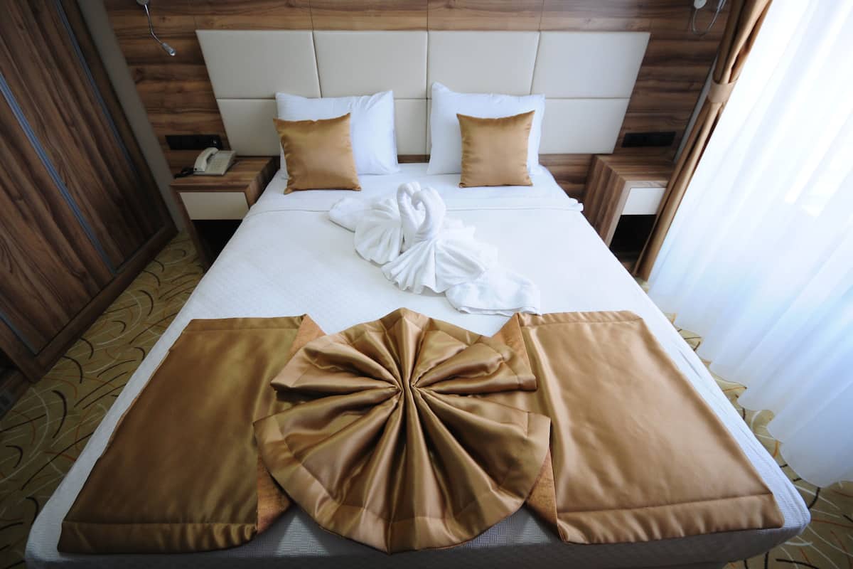A hotel room with a neatly made bed, a wooden headboard, a bedside table with a phone, and a nightstand with a lamp.