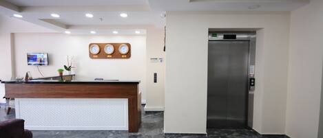 A reception area with a wooden desk, a TV mounted on the wall, a clock, and a modern elevator.