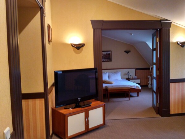 Quarto de hotel com televisão sobre um armário de madeira, cama com roupa branca e uma pequena mesa de cabeceira.