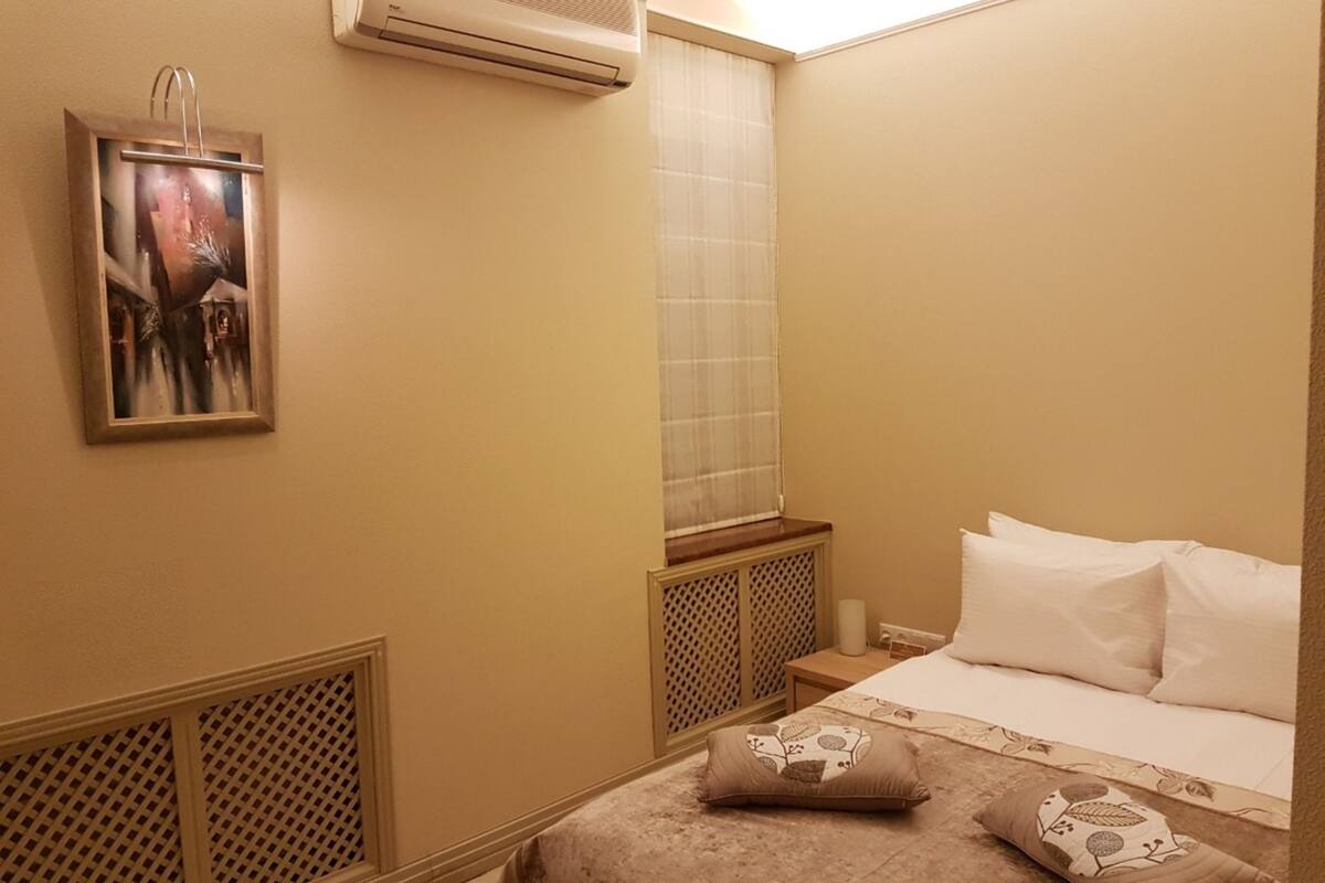 A bedroom with a bed, pillows, a window with a curtain, an air conditioning unit, and a painting on the wall.