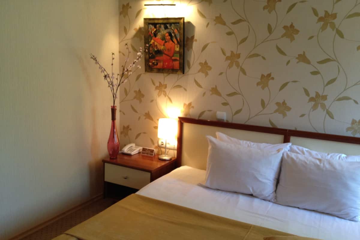 A hotel room with a bed, a nightstand with a lamp, a vase with branches, and a painting on the wall.