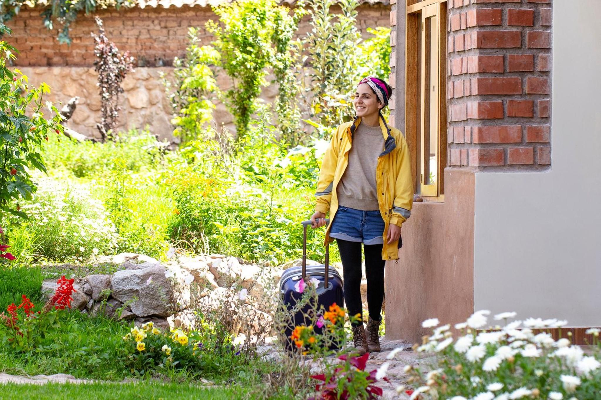 Una persona de pie junto a una maleta en un jardín con un muro de ladrillo y follaje.