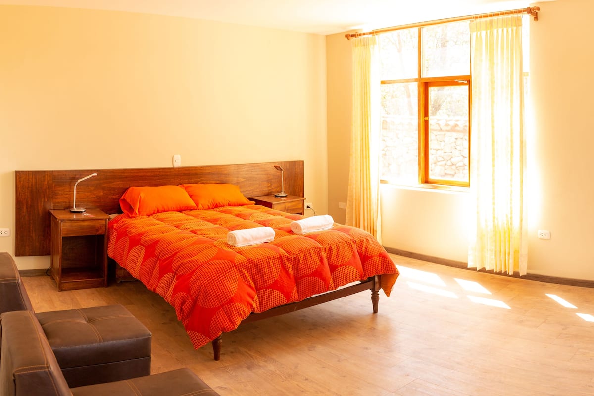 Un dormitorio con cama de madera, una mesita de noche, una ventana con cortinas y una puerta que da al baño.