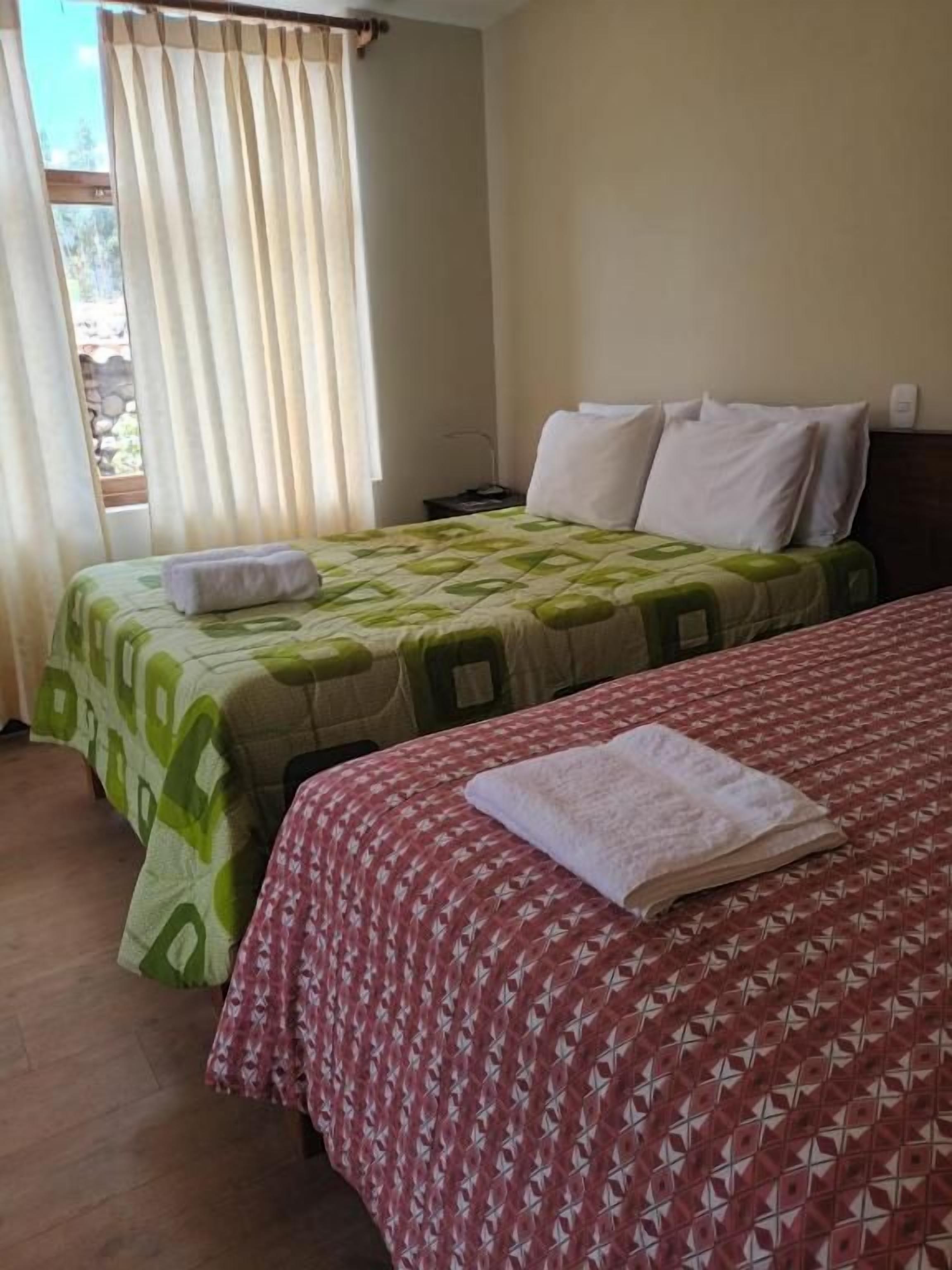 A room with two beds, one with a green and white patterned bedspread and the other with a red and white patterned bedspread. There is a window with curtains, a bedside table with a lamp, and a wall-mounted light switch.
