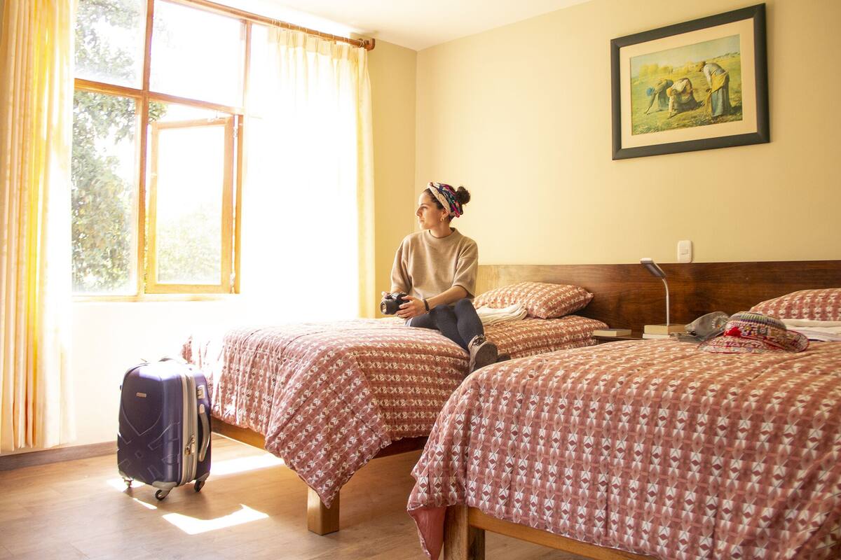 Una habitación de hotel con dos camas, un ventanal con cortinas, un cuadro en la pared y una maleta en el suelo.